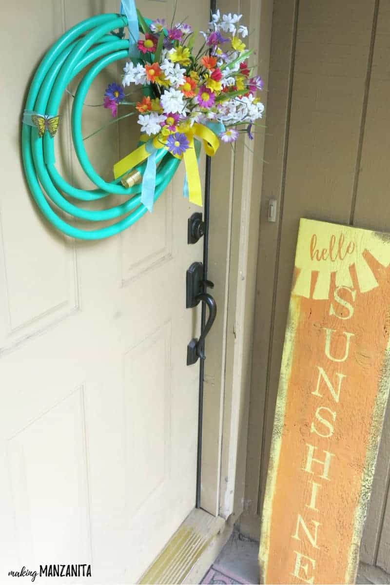 A colorful summer garden hose wreath with a faux flower bouquet hangs on a tan front door, next to a hello sunshine front porch sign.