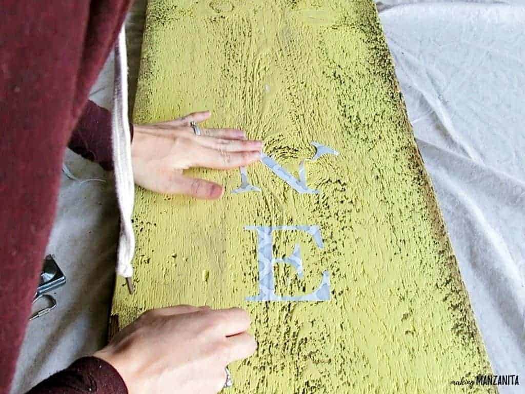 A woman is starting to apply the cut-out vinyl letters to the yellow painted wood sign, starting with the N and E.