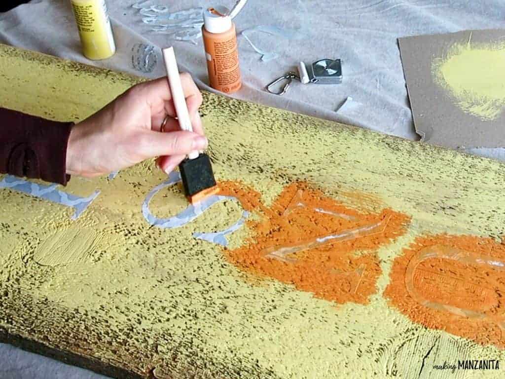 A woman uses a sponge brush to paint over the vinyl letters using orange paint.