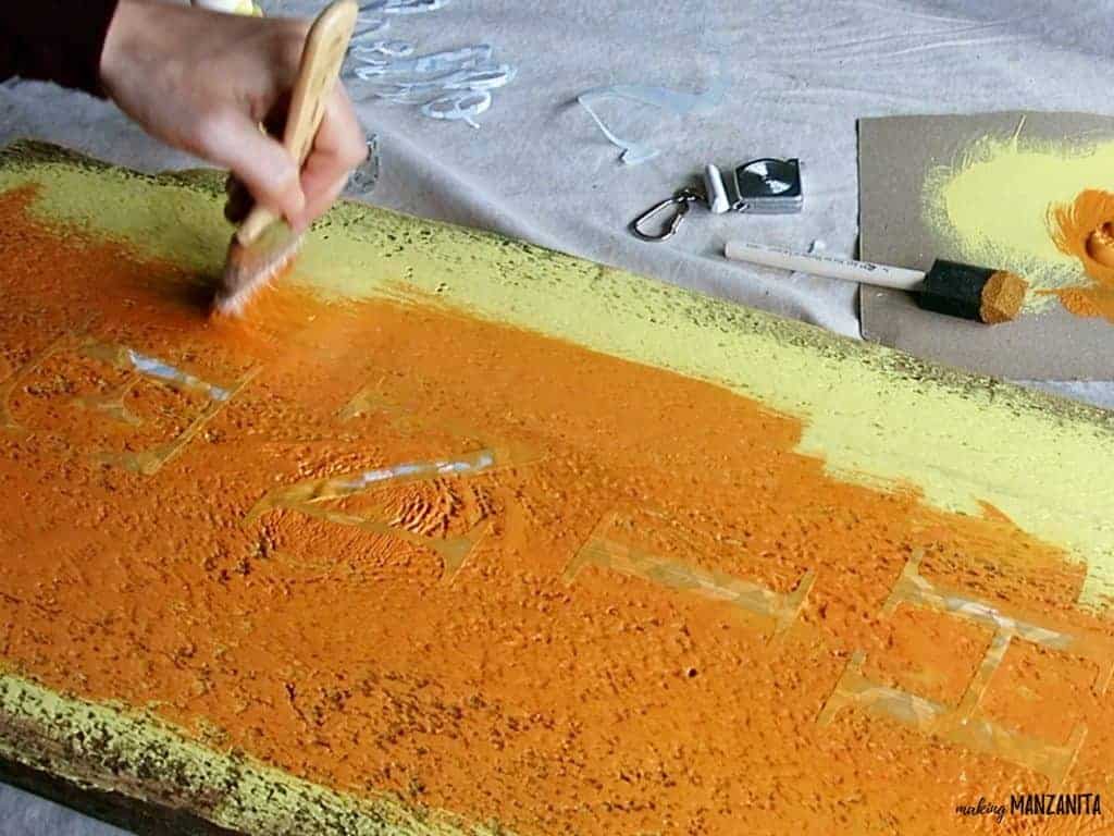 A woman paints a coat of orange paint over the yellow painted wood sign and vinyl letter stencils.