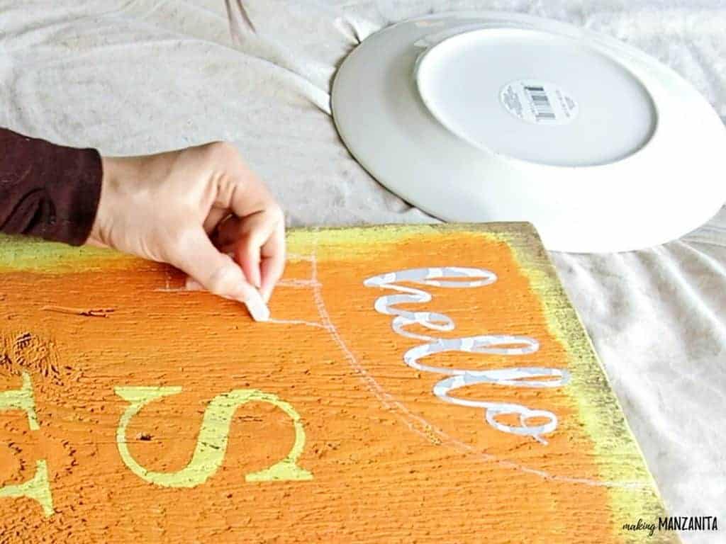 A woman uses a piece of white chalk to draw a sun shape and sun rays on the top of the painted wood sign. The word "hello" is applied in a vinyl letter stencil.