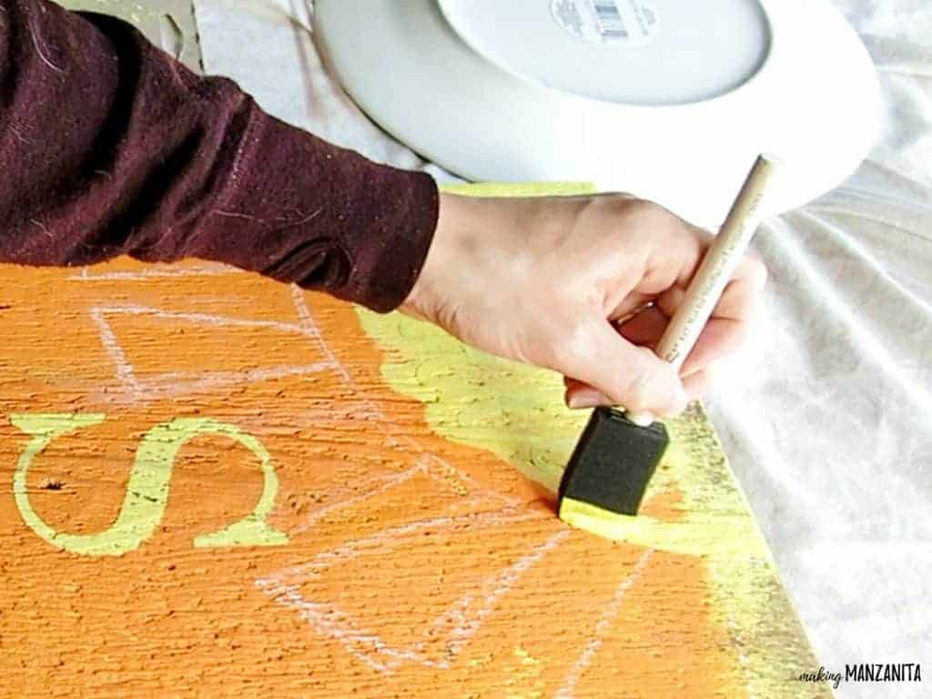 A woman uses a sponge brush to paint a yellow sun shape at the top of the wood sign, over the vinyl stencil.