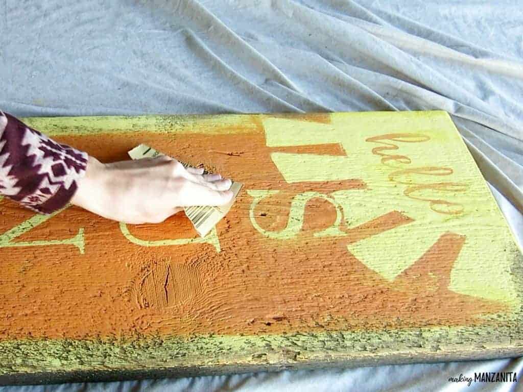 A woman lightly sands the surface of the finished hello sunshine sign with a piece of sandpaper, giving it a distressed look.