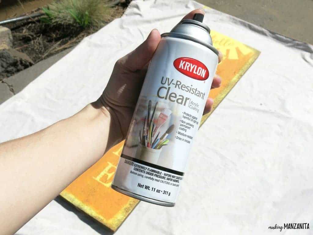 A woman holds a can of Krylon's UV-Resistant Clear Coating with the hello sunshine wood sign sitting on a tarp in the background.