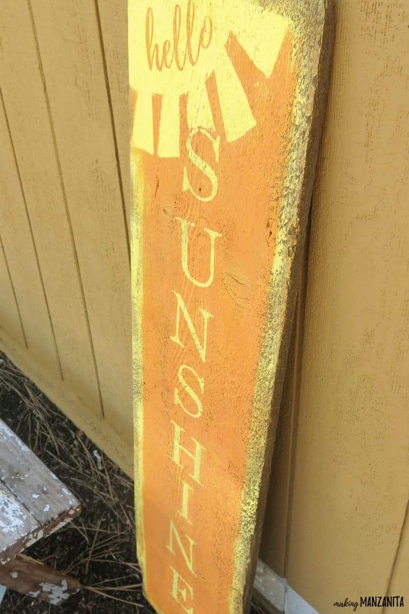 A look at the completed Hello Sunshine wood sign, painted with yellow and orange paint, sitting against a gold painted exterior wall.