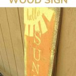 Shows a yellow and orange sign that says hello sunshine standing up against a brown wall outside that has text overlay that says hello sunshine wood sign at top