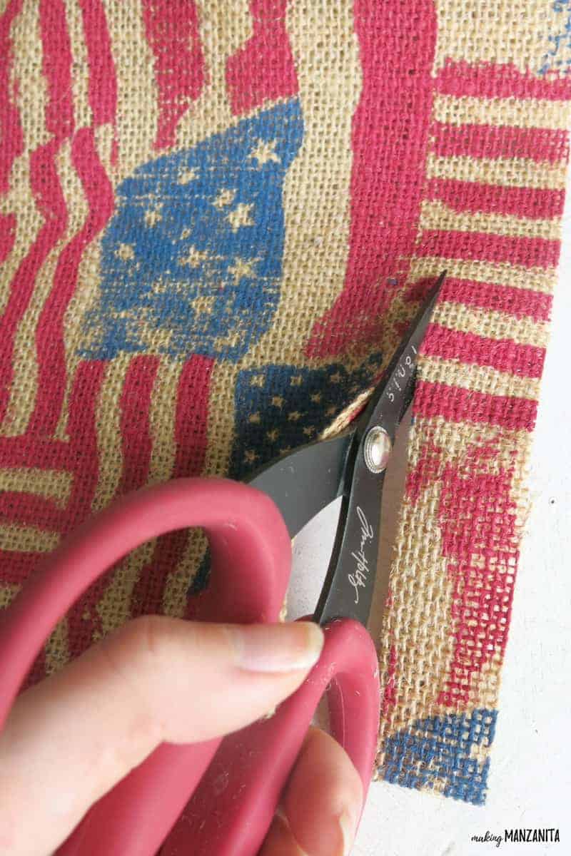 Cutting strips of American flag printed burlap to make a 4th of July flag ribbon window hanging