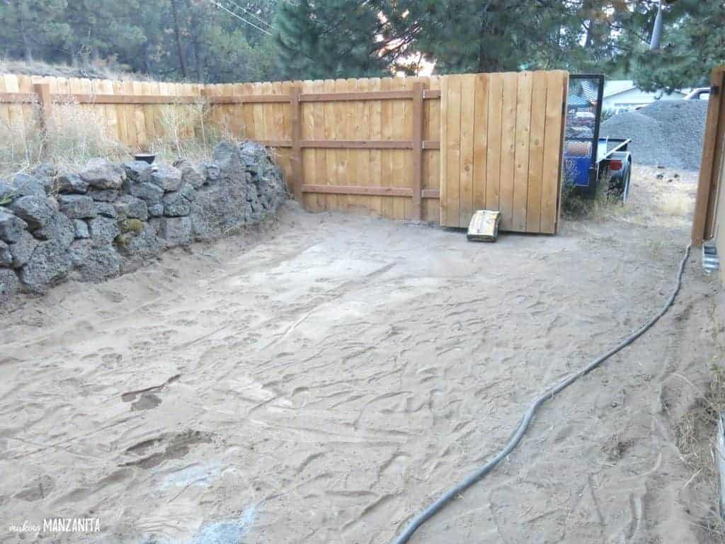 Flat and empty dirt ground  surrounded with rock bed and wood fence and open gate before wood garden beds were installed