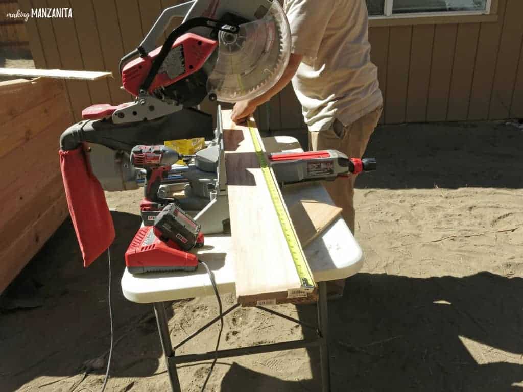 Man measuring the cedar boards on the table beside the miter saw before cutting down to size to install on the raised garden beds in backyard 