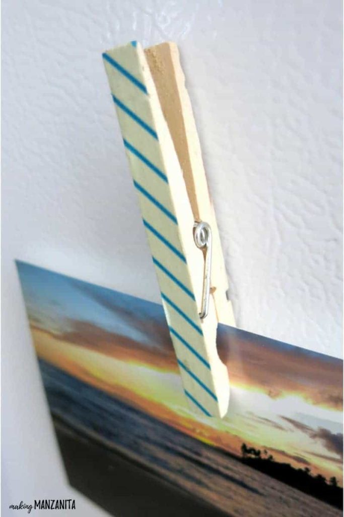 A close-up image of a clothespin magnet holding a picture of a sunset to a white surface. The wooden clothespin is covered with a white and blue striped washi tape.