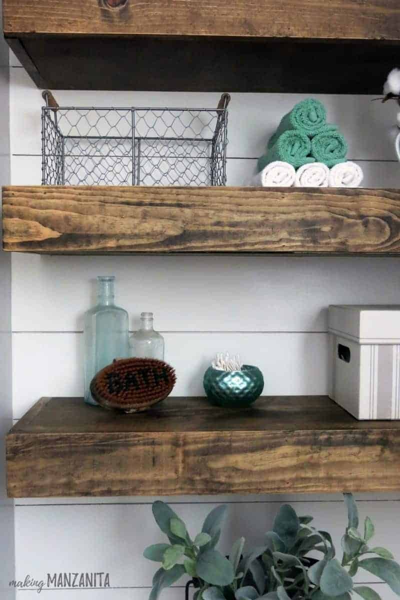 Floating wood shelves above the toilet in our farmhouse style bathroom are decorated with little trinkets like vintage glass bottles, wire baskets, and rolled tea towels.