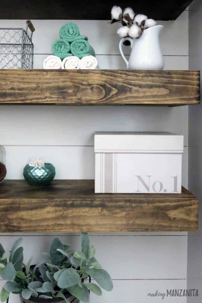 Floating shelves above the toilet in our farmhouse master bathroom, decorated with trinkets like tea towels, bowls, and a white tissue box.