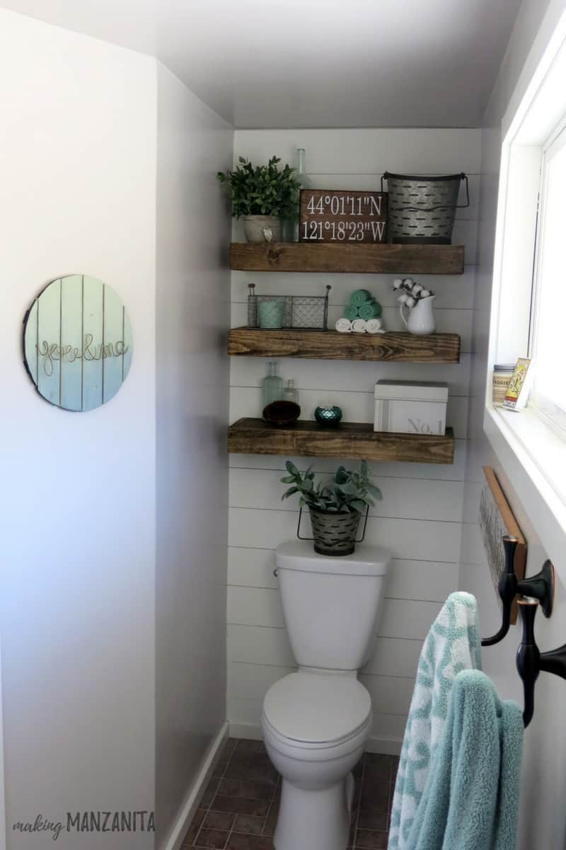 A view of the toilet alcove in our farmhouse master bathroom; floating shelves are mounted on a shiplap wall behind the toilet, towel hooks hang under the window, and a vintage round wood sign hangs on the wall.