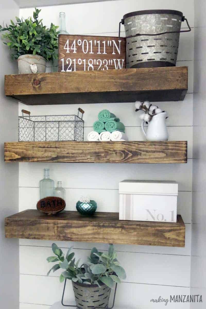 Another look at the floating shelves mounted above the toilet in our farmhouse master bathroom, decorated with fun trinkets like wire baskets, tea towels, a wood coordinates sign and more.