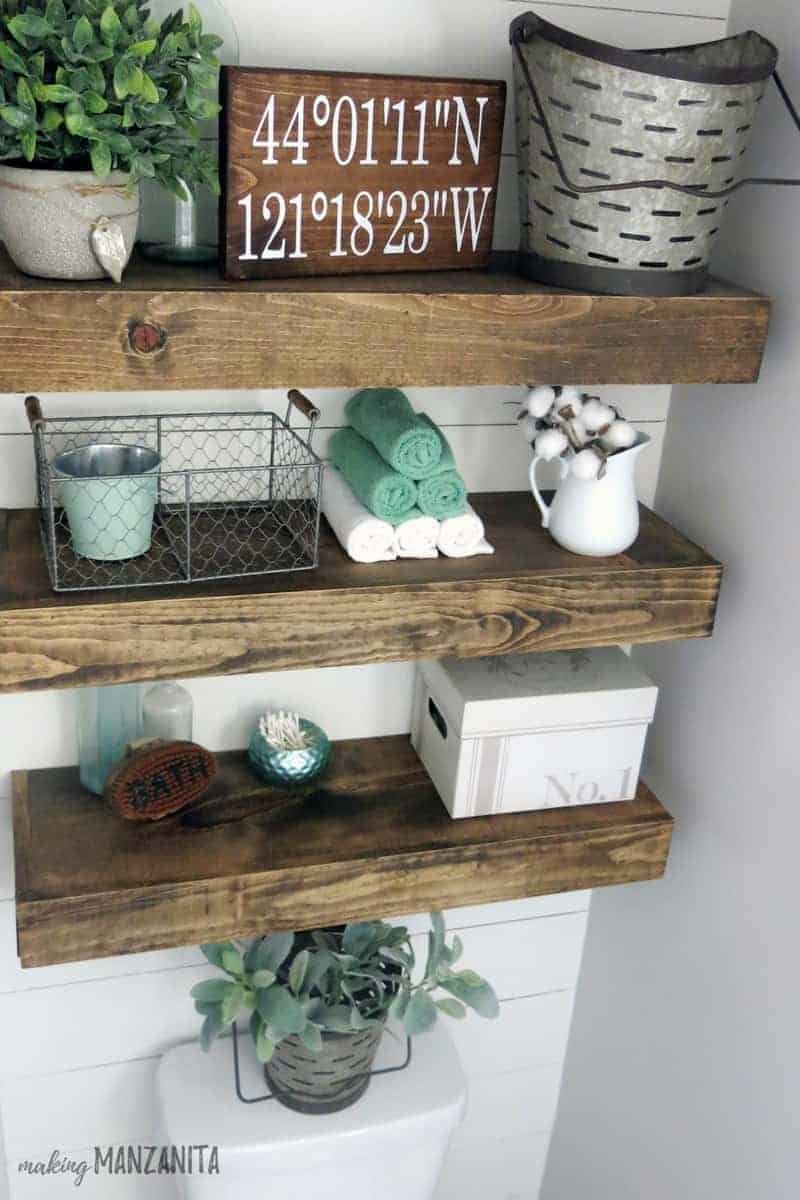 We installed floating wood shelves over the toilet in our farmhouse master bathroom. The shelves are decorated with small trinkets like towels, baskets, plants, a coordinates sign, and more.