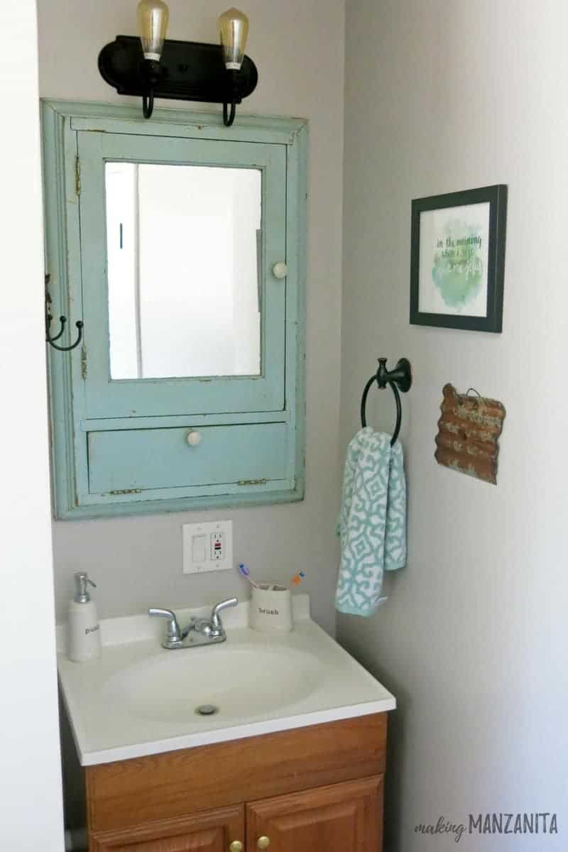 The sink and mirror vanity in our farmhouse master bathroom; the medicine cabinet and mirror is painted a weathered mint green, brass towel rings and hooks are mounted to the walls. 