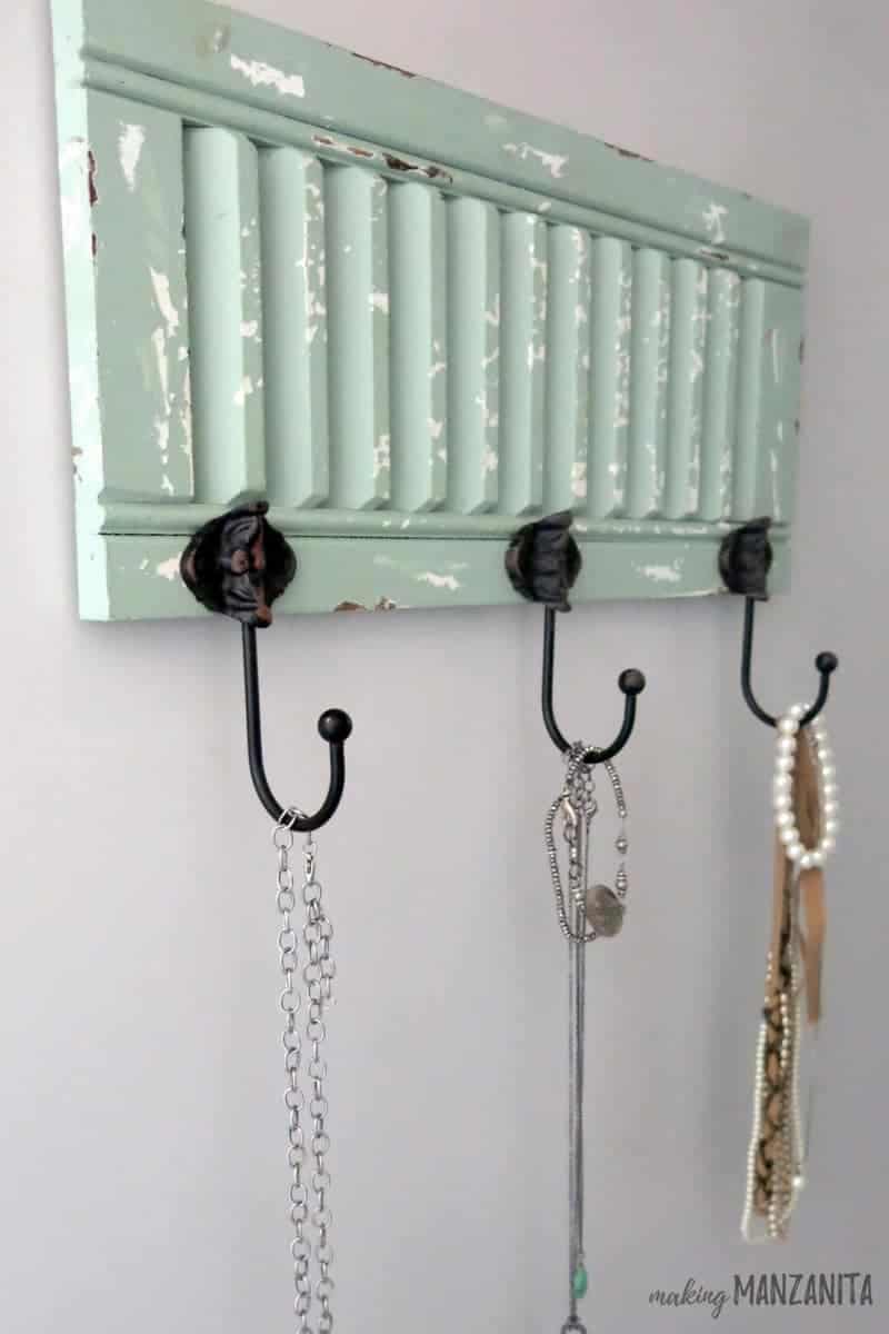 This DIY jewelry holder was made with an old vintage shutter painted with chipped mint green paint, and has three metal hooks attached to hold bracelets, necklaces, and other jewelry.