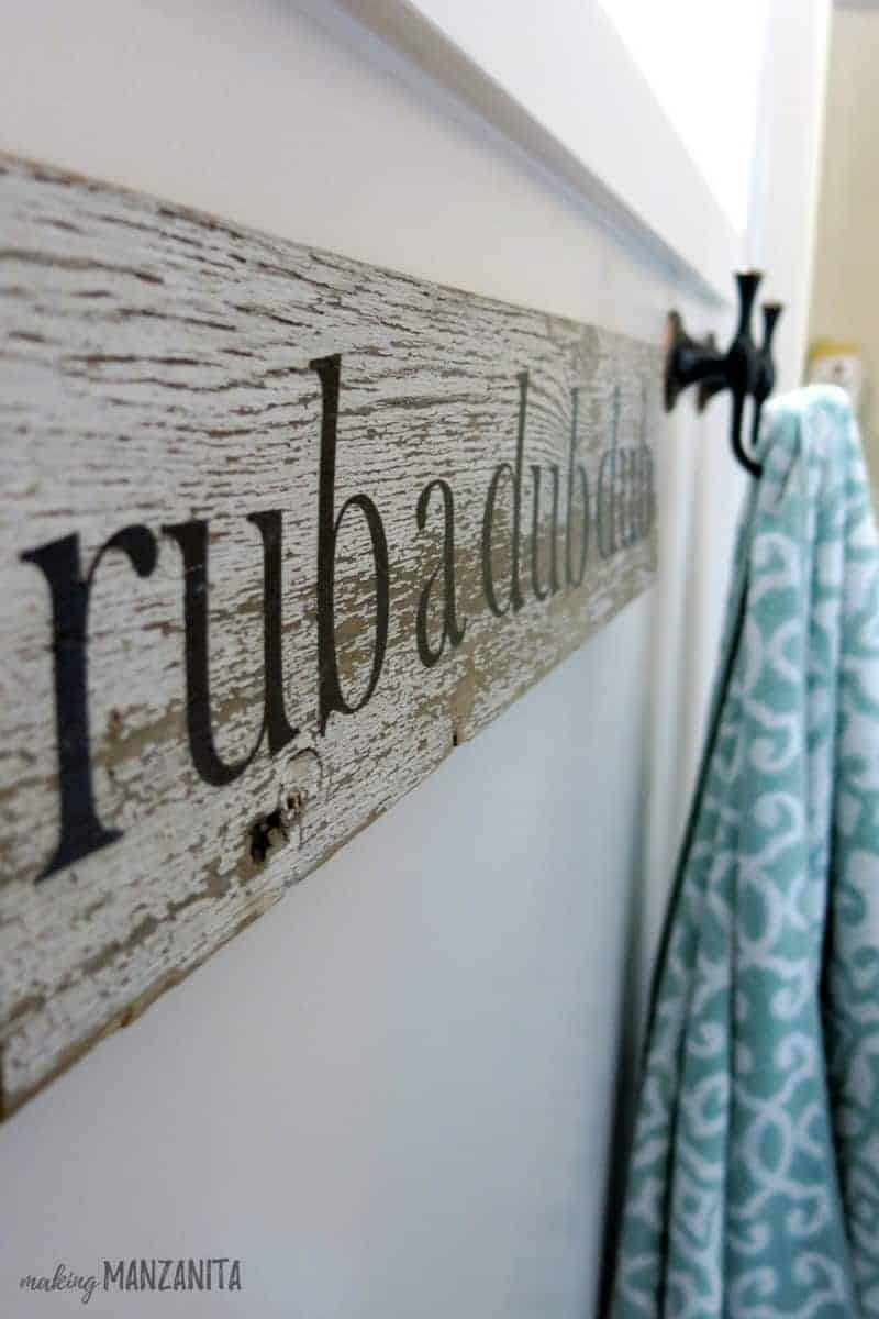 A close-up angled look at a rustic wood sign that says "rubadubdub" and hangs on the wall next to a set of towel hooks.