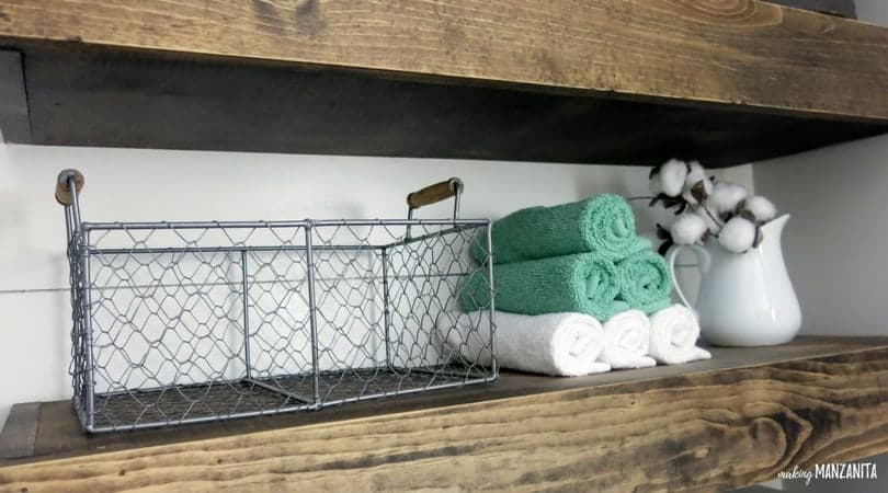 A chicken wire basket, rolled up tea towels, and a vase with cotton blooms sits on the floating shelves above the toilet.