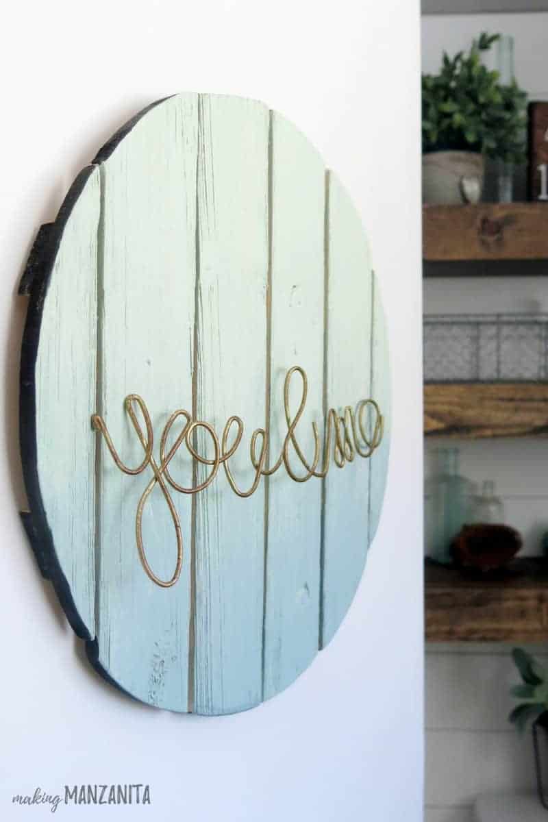 A blue and teal ombre painted sign has metal wire twisted to read "you & me" hangs on a white wall.
