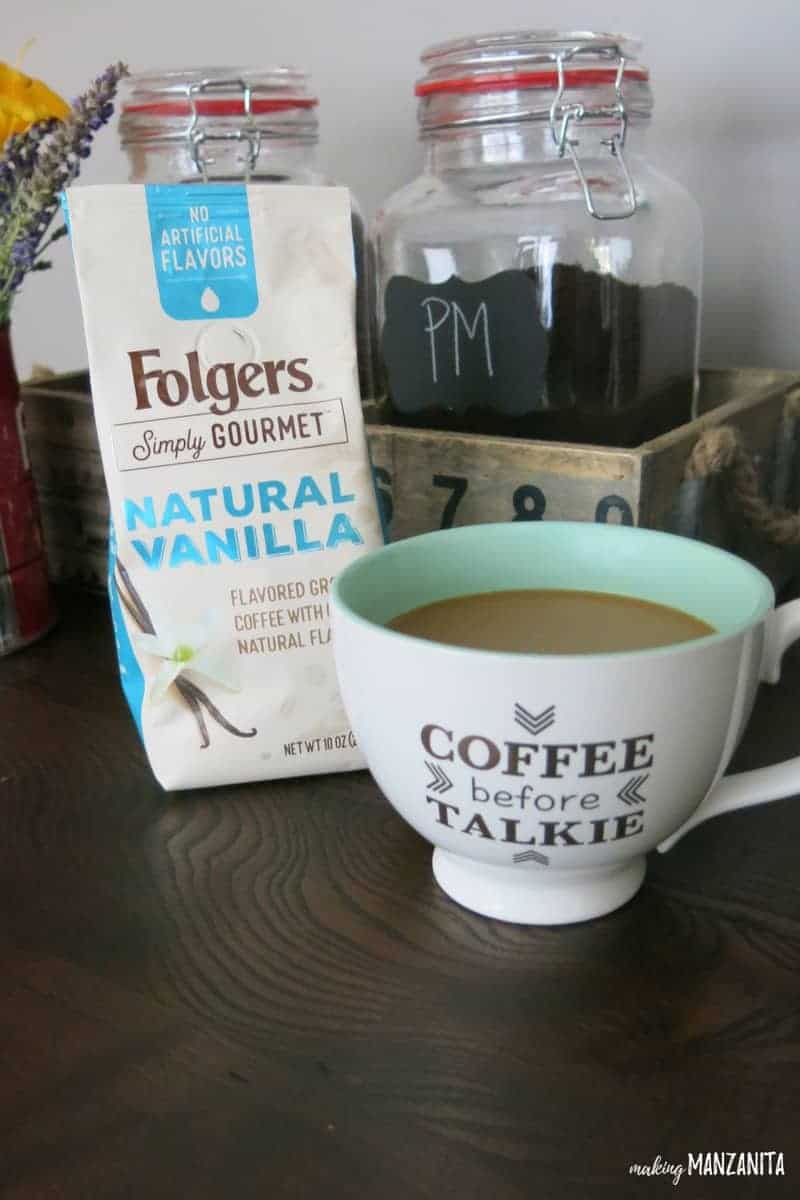 We loved this Folgers simply gourmet natural vanilla. It looks great next to a white and teal coffee cup that says coffee before talkie on a wood table with some glass containers behind at our coffee station in our dining room