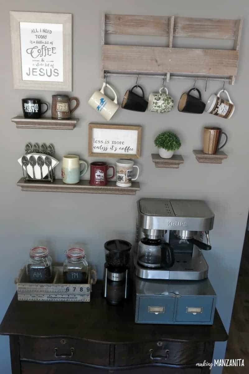 We used a vintage dresser to create a DIY coffee station with coffee beans, a coffee grinder, and a coffee maker on top of a metal drawer. On the wall, there's a wood mug hanger floating shelves holding mugs and a farmhouse sign
