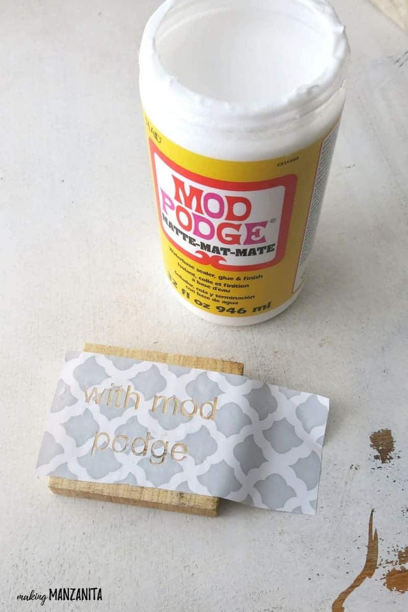 Bottle of Mod Podge sitting next to piece of wood with stencil that says With Mod Podge