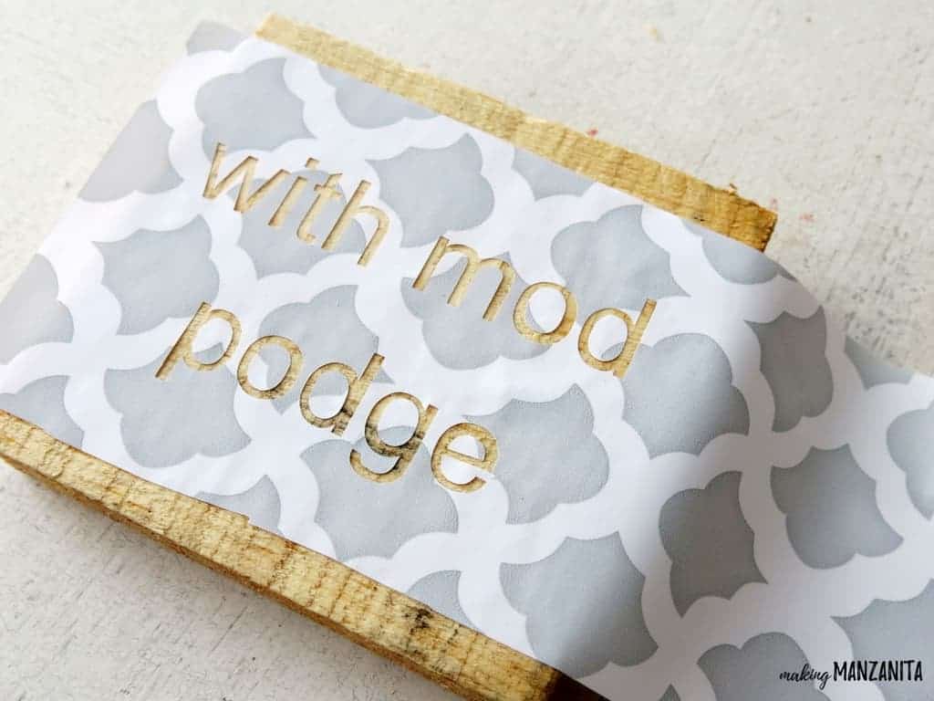 Piece of wood with stencil attached that says with mod podge
