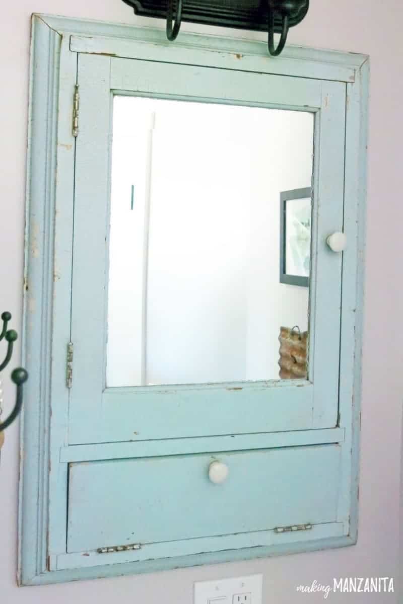 A vintage medicine cabinet hanging on a bathroom wall - the medicine cabinet is painted with chipped mint green paint, has a mirror and two small storage doors.