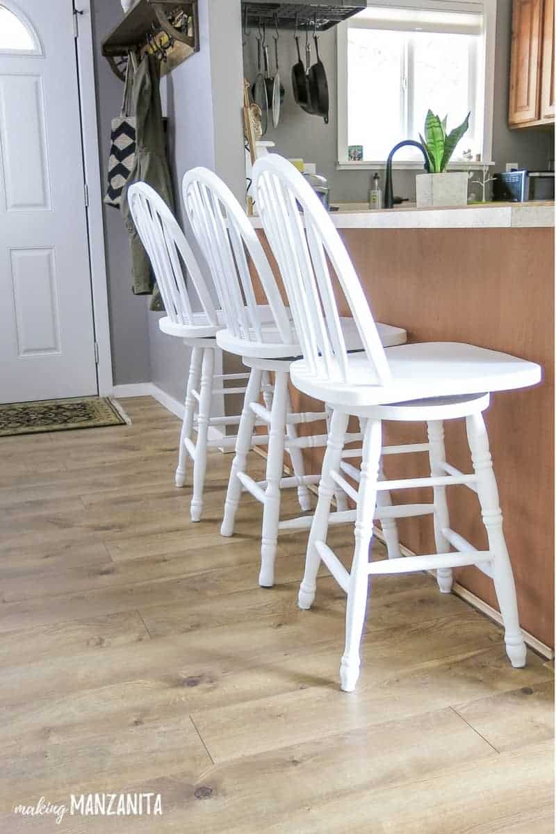 Three white chalk painted bar stools sit lined up at a breakfast bar.