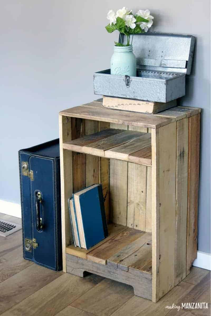 The completed rustic pallet wood side table, decorated with vintage books, a metal box and mason jar vase with flowers, and a navy blue vintage suitcase.