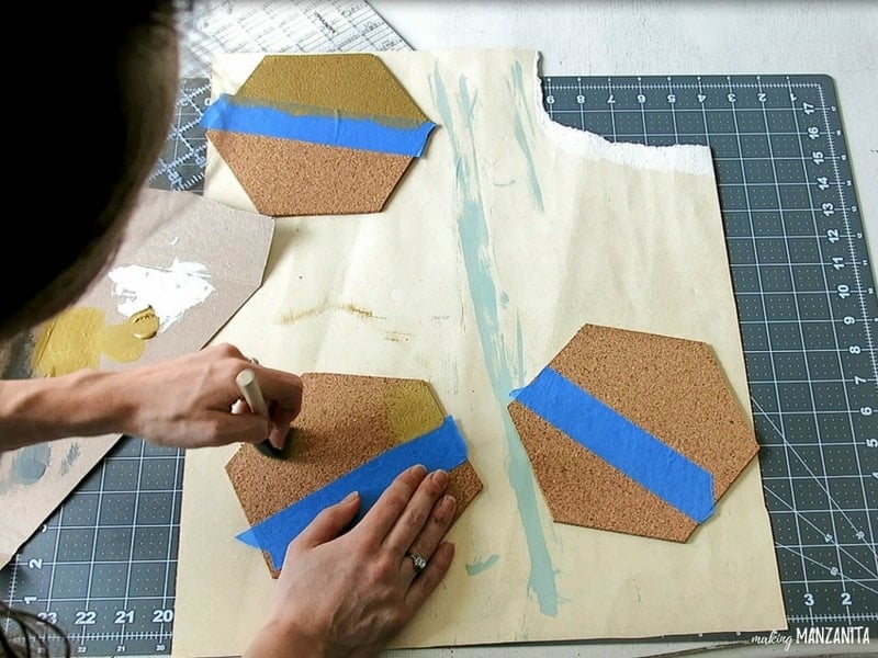 Woman holding a paint brush painting a hexagon cork board with gold acrylic paint while holding the other