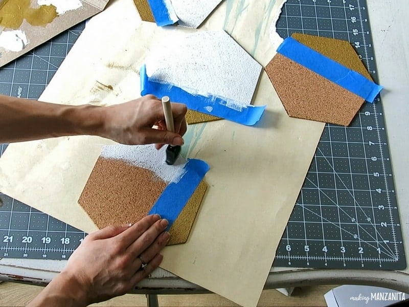 Woman holding and painting one hexagon cork board beside the two other hexagon cork board on top of the paper and cutting mat