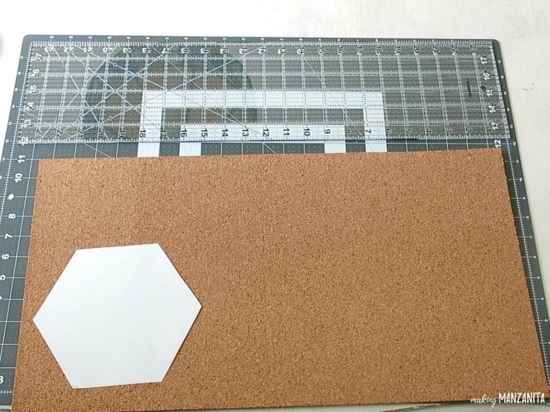 Hexagon printed and cut out of white copy paper placed on top of cork sheet and cutting pad with measurements before cutting cork to make DIY hexagon memo boards