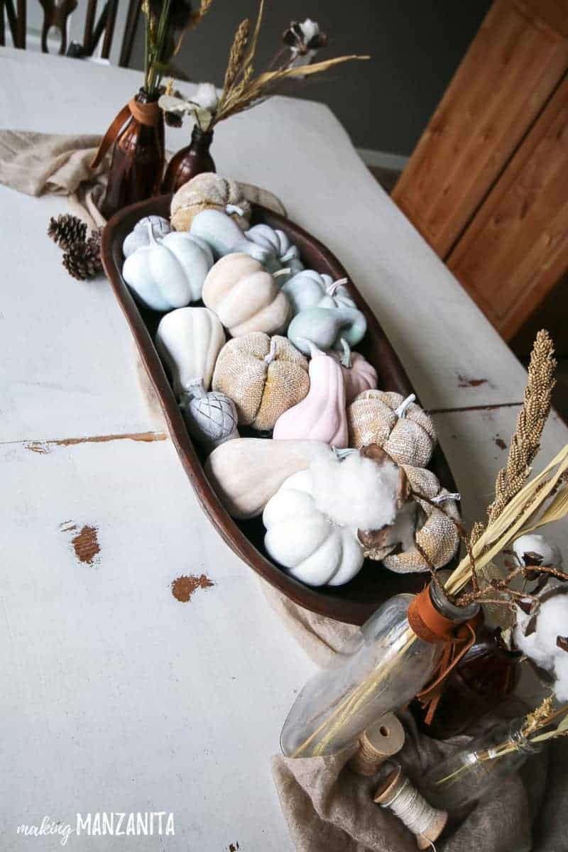 Wooden bowl holding pastel colored painted mini pumpkins and goards to decorate table for fall next to vintage bottles with dried wheat and leather