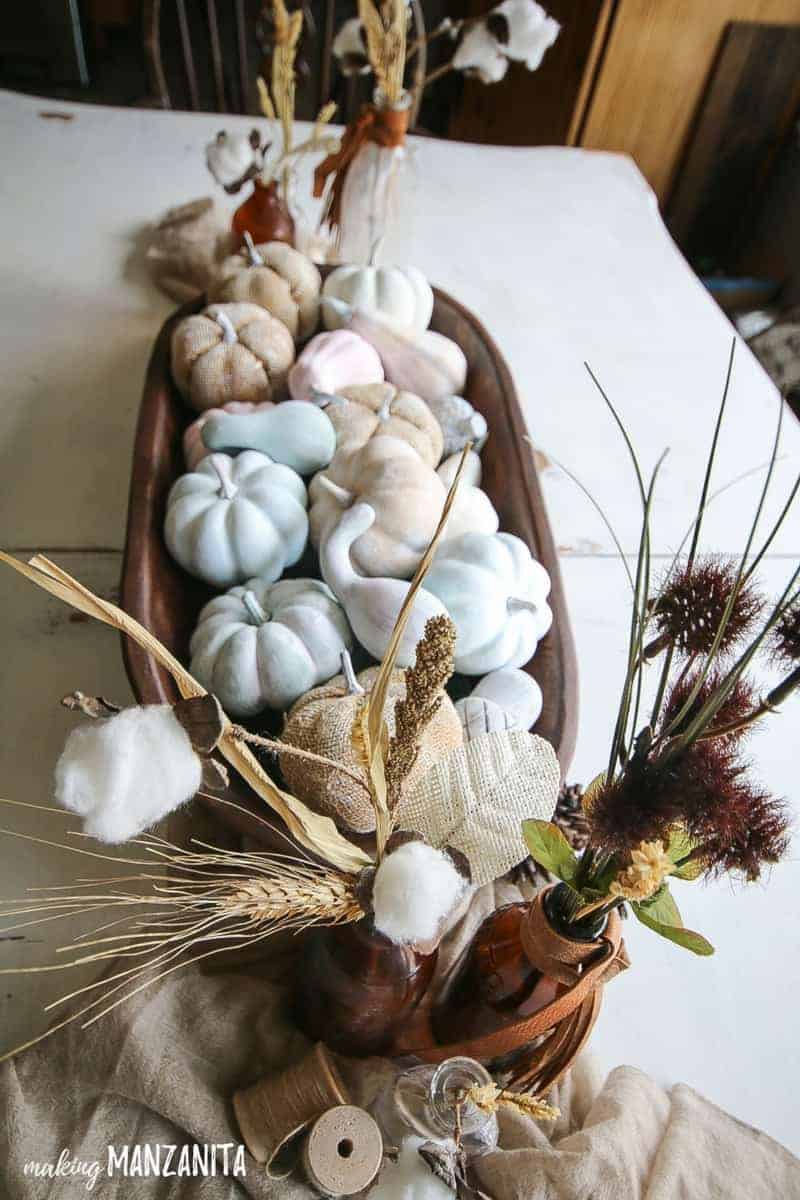 Side view of pastel farmhouse fall table decorations with farmhouse style that incorporate vintage bottles, mini painted pumpkins, wooden dough bowl, tan fabric on a white farmhouse table