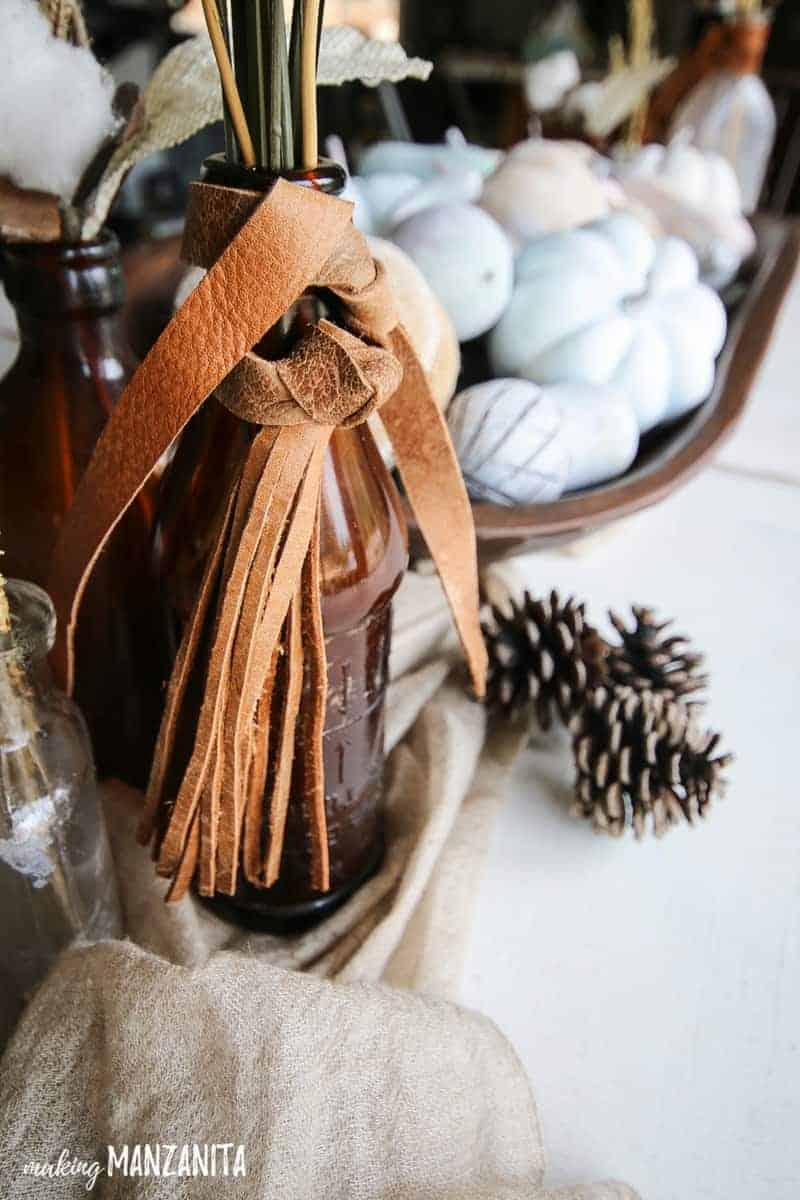 Vintage amber glass bottle with leather tied around the top and pine cones laying nearby on white table with painted mini pumpkins in background