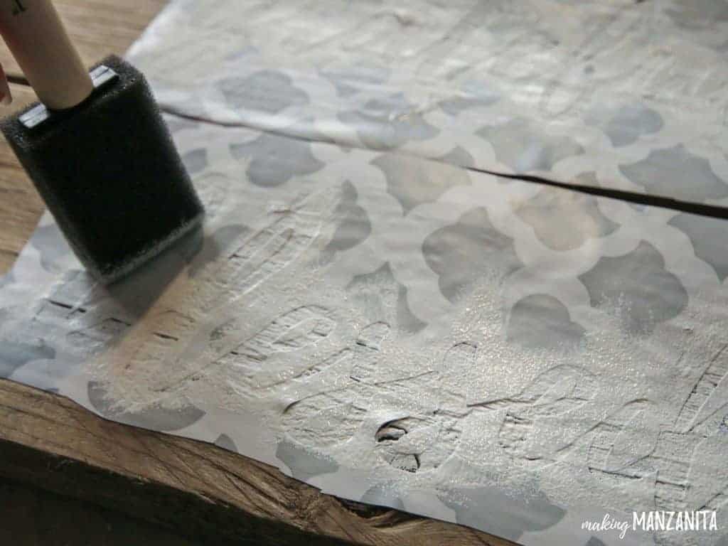A woman uses stencils, white paint, and a sponge brush to paint the word "blessed" in cursive text over the surface of a wooden pallet board.