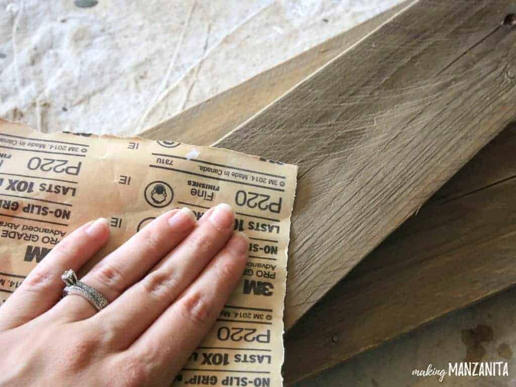 A woman uses a square of sandpaper to sand down pieces of pallet wood.