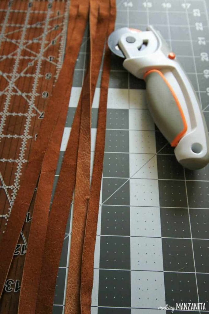 A rotary cutter sits next to thin strips of caramel colored leather.