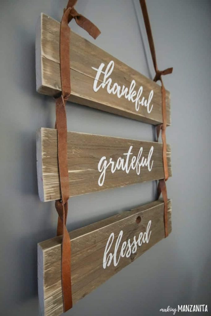 A closeup side view of a "thankful, grateful, blessed" wood pallet sign hanging on a gray wall. The pallets are tied together with brown leather straps.