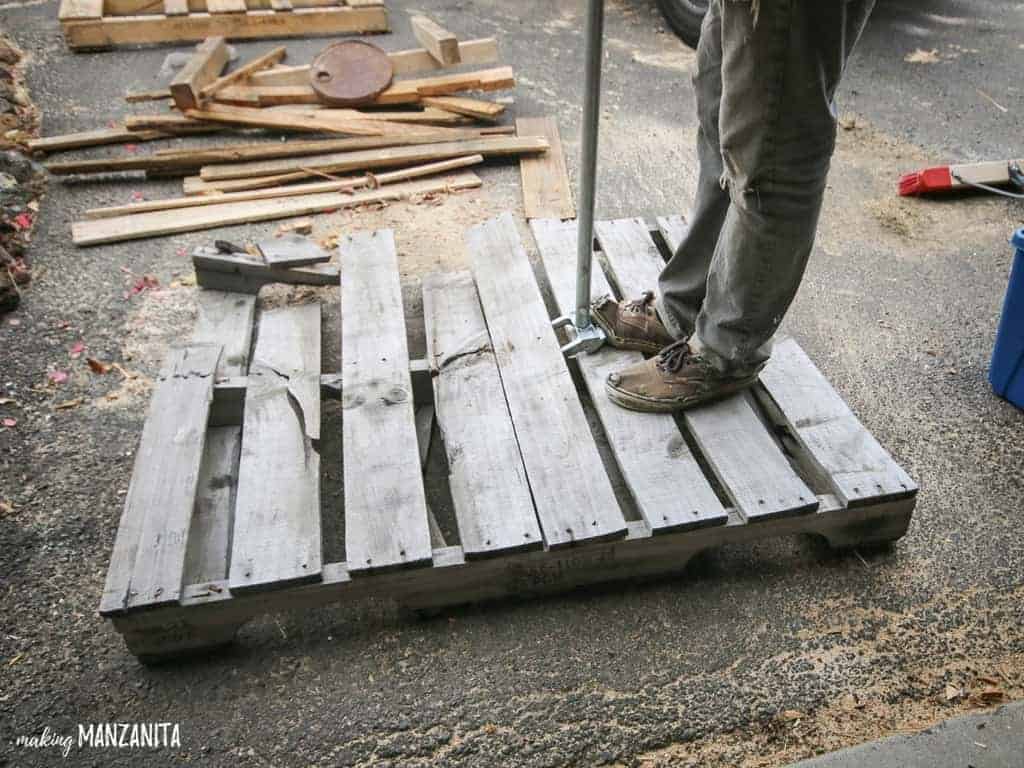 A man standing on a recycled wooden pallet, using a pallet jack to rip apart the boards of the pallet.