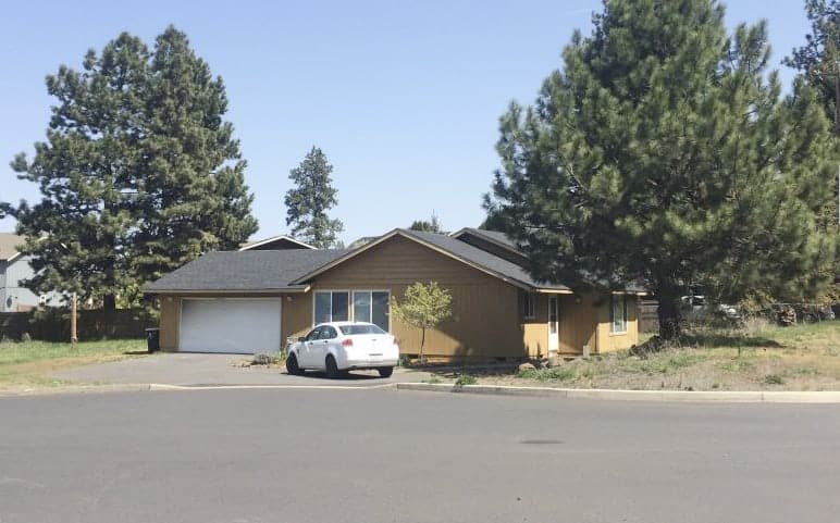 A wide view of our ranch style home with a tan exterior, surrounded by trees and a white car in the driveway