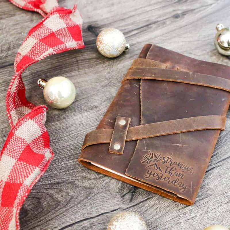 Leather wrap journal with quote engraged in corner that says stronger than yesterday on wood background with gold mini Christmas ornaments and red plaid ribbon laying next to it