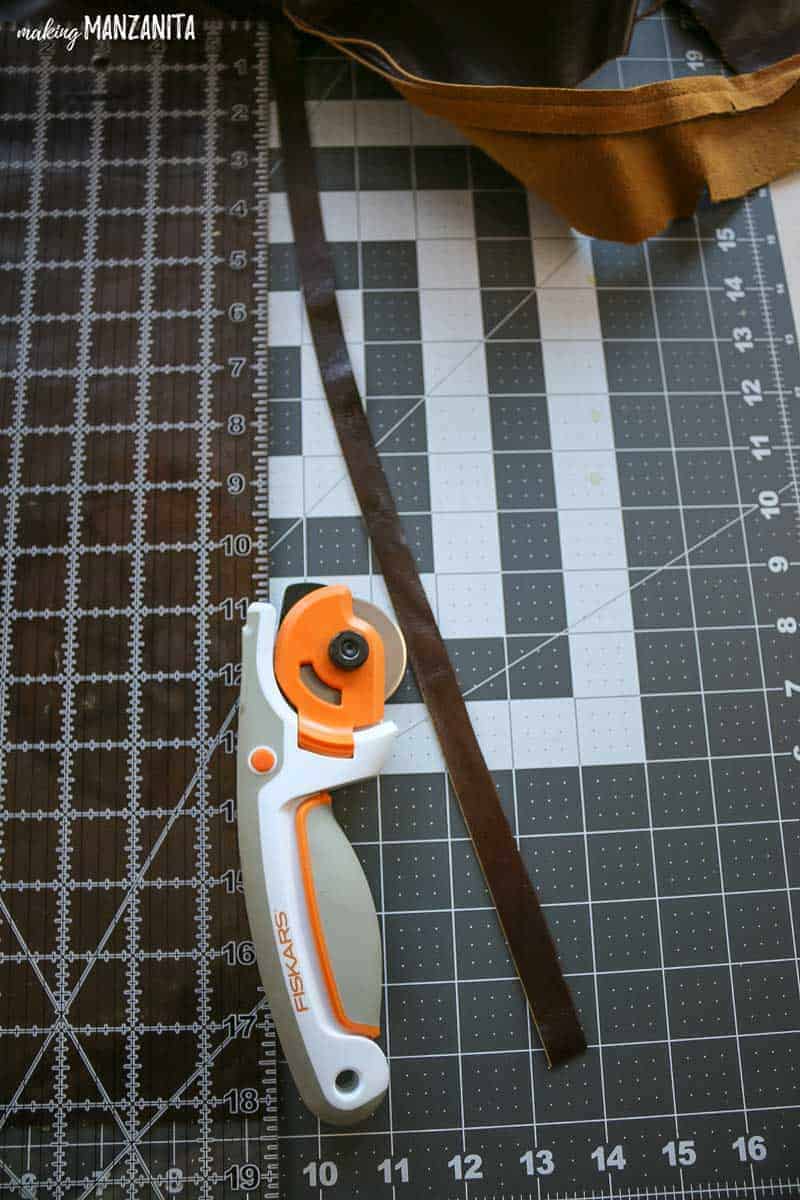 Large piece of brown leather laying on a cutting mat with a long 1 inch wide strip cut in the leather with a rotary cutter and acrylic ruler laying on table