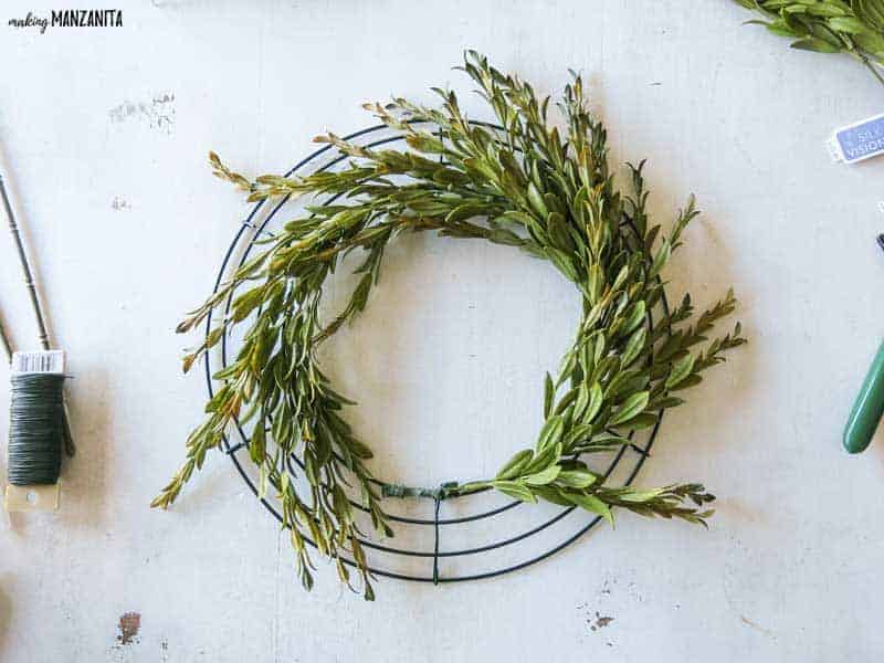 The wire wreath frame is filling up with faux greenery to create a farmhouse wreath