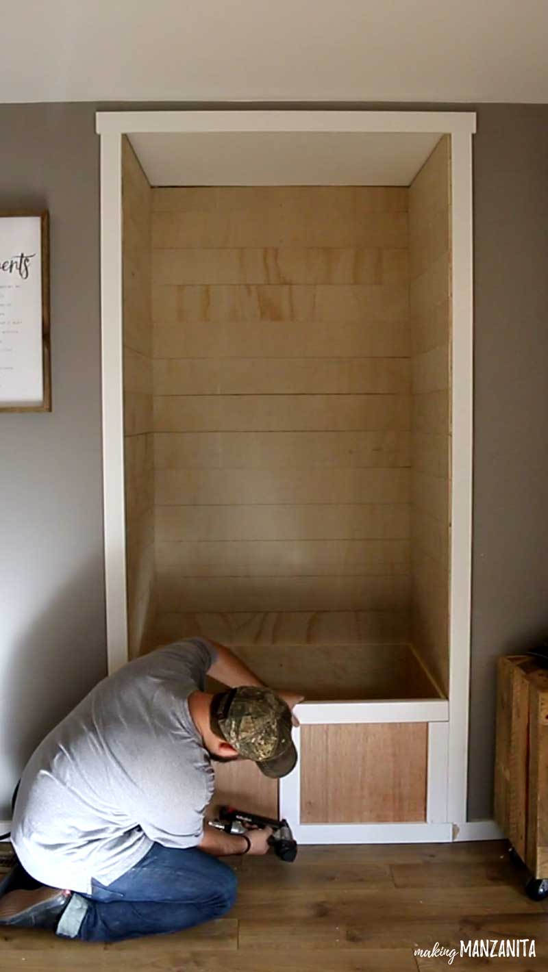 Logan is using a nail gun to install the white trim frame on the front of the storage box/bench in the living room reading nook.