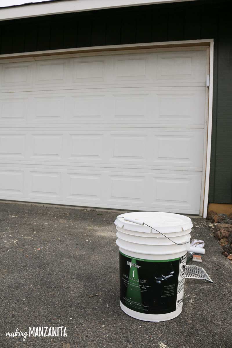 A large bucket of Behr Marquee Exterior paint sitting on the driveway in front of the garage.