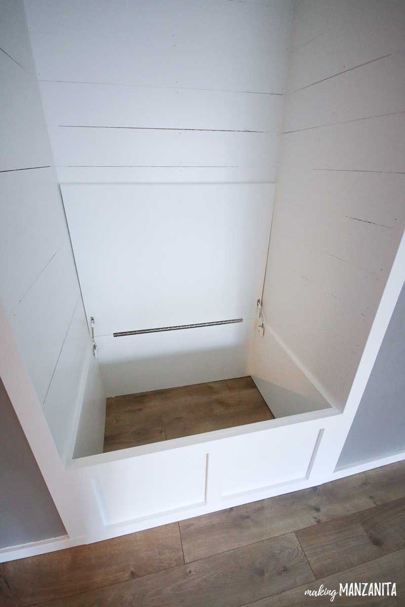 A look inside our in-progress reading nook - the shiplap walls and storage bench have all been painted white, and the bench lid has been installed - and sits open with hinges exposed.