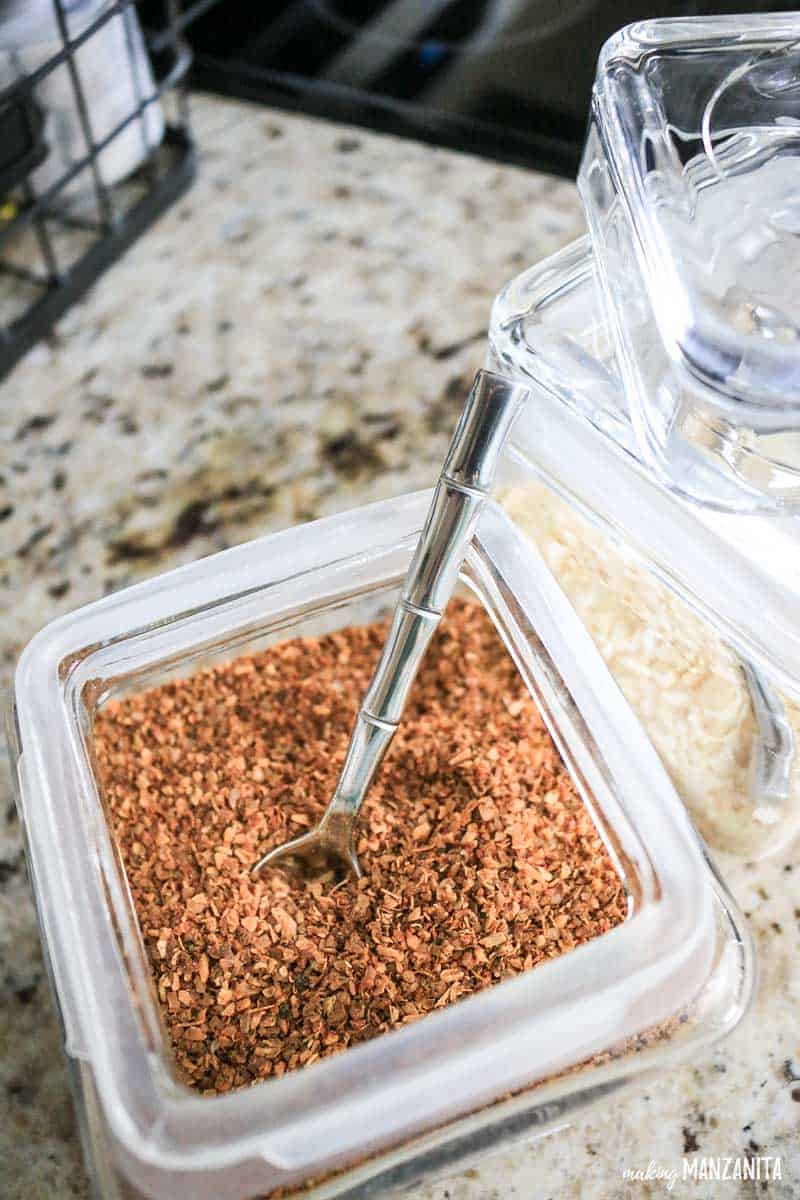 A large stackable spice container sitting on the kitchen counter, open with a spoon dipped into the mesquite spice.